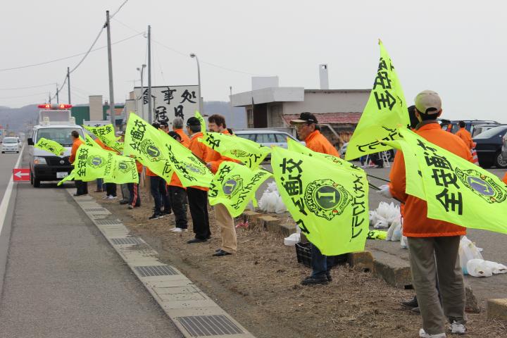 風の強い中交通安全の旗を振り安全運転を呼びかけるLメンバー